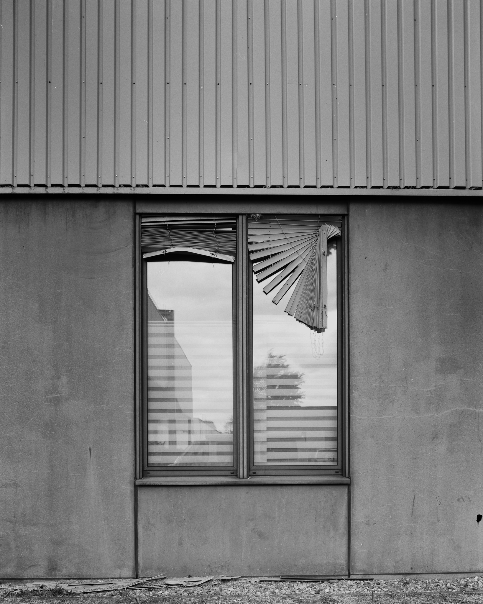 A wall with a window, trees and a house reflecting in it. There are blinds on top of each window half, one of them broken hanging down in a odd angle.