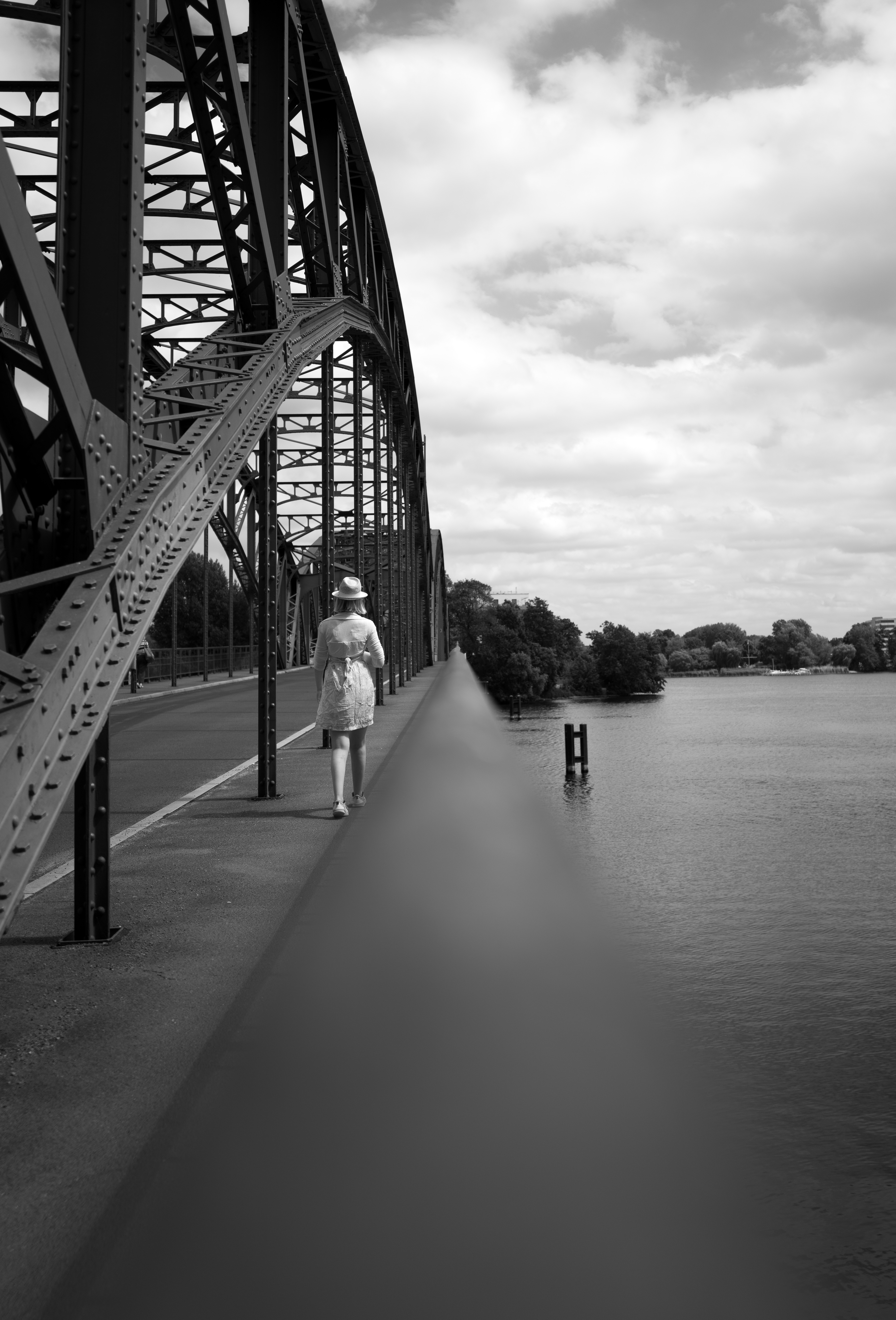 A woman walking down a bridge, the matal construction of the bridge on the left side of the frame balanced by the nature and sky on the right.